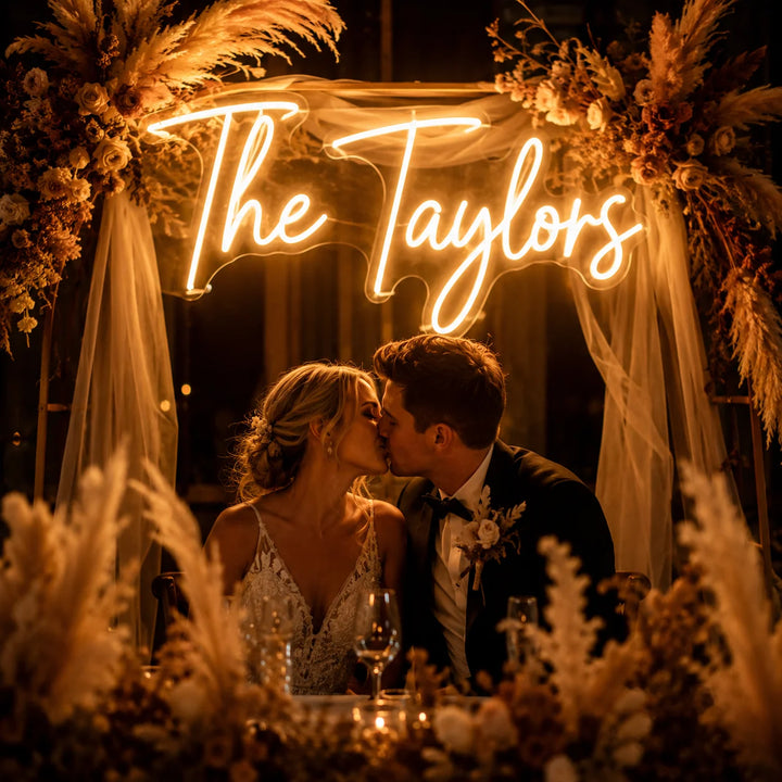 Wedding neon sign featuring “The Taylors” custom surname design with warm white LED lighting on clear acrylic backing for romantic wedding reception backdrop decor.