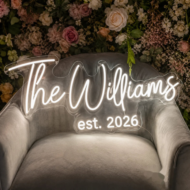 Custom wedding LED neon sign with “The Williams est. 2026” on clear acrylic backing, displayed on a velvet chair with floral wedding backdrop decoration.