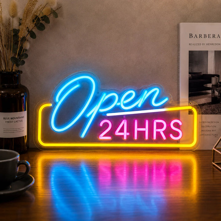Colorful “Open 24HRS” LED neon sign glowing in blue, pink, and yellow on a wooden table with café decor and warm ambient lighting.