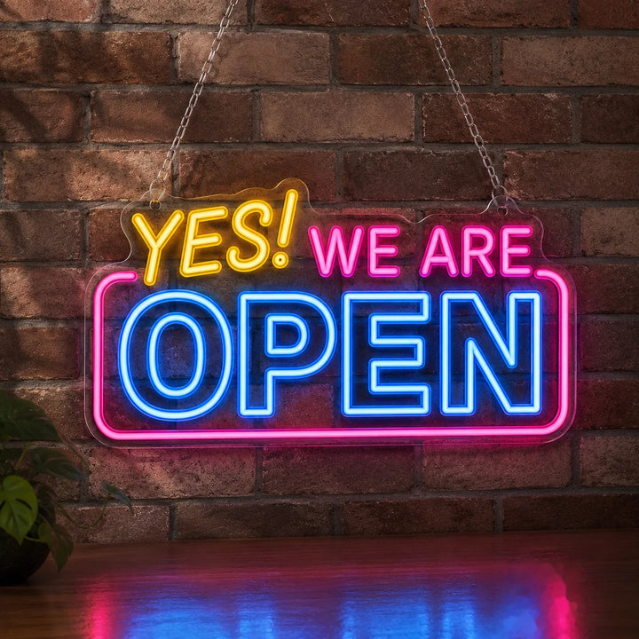 Bright “YES! WE ARE OPEN” neon wall sign with vibrant blue and pink LED lighting displayed against a rustic brick wall in a modern café or storefront setting.