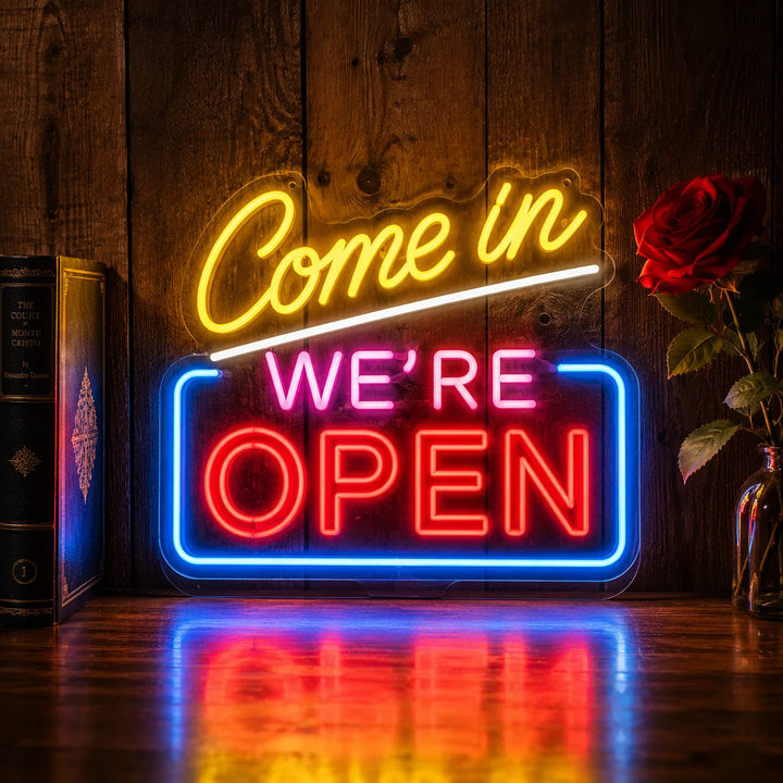Bright “Come In We’re Open” neon wall sign glowing in yellow, pink, red, and blue colors against a wooden background with rose decoration and warm ambient lighting.