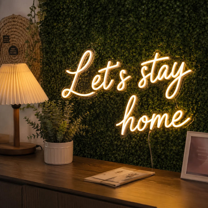 Decorative warm white neon wall sign reading “Let’s Stay Home” with greenery backdrop, table lamp, and cozy home interior styling.