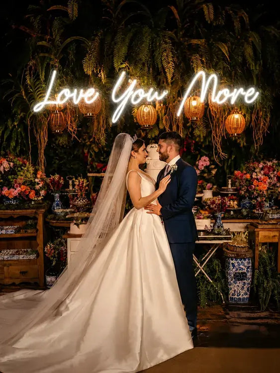 A bride in a white satin wedding gown with a long veil and a groom in a navy suit embrace under a glowing 'Love You More' neon sign, set against a tropical night backdrop with hanging lanterns and floral arrangements.