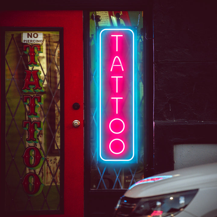 Vertical pink and blue neon LED sign spelling TATTOO displayed in a shop window.