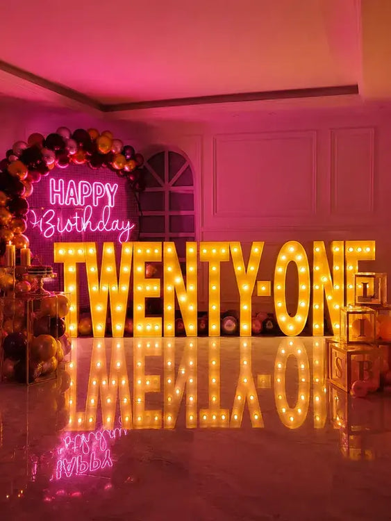 A party setup in a room with a large illuminated marquee sign spelling 'TWENTY-ONE' in warm yellow lights, a pink neon 'HAPPY Birthday' sign, and a balloon garland on the left.